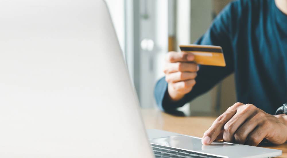 Zoomed in image of a person’s hands. One hand is on a laptop. The other hand is holding a credit card.