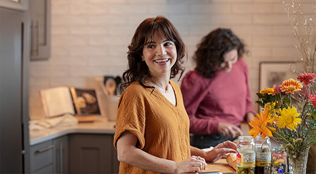 Two people preparing food in a kitchen with a cutting board, jars, and flowers on the counter.